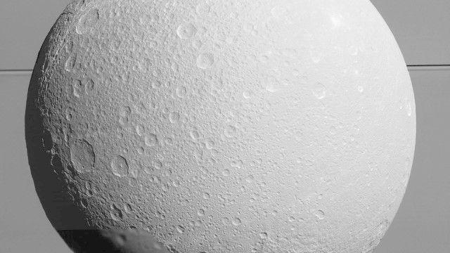 Dione vista pela Cassini durante a aproximação de segunda-feira, no quinto e último sobrevoo da sonda por esta lua de Saturno, ao fundo com seus anéis, em missão que já dura mais de dez anos
