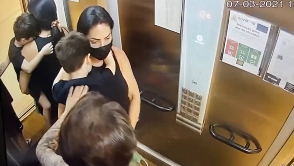 Henry no colo da mãe enquanto Dr. Jairinho o acaricia, no elevador do prédio em que moram